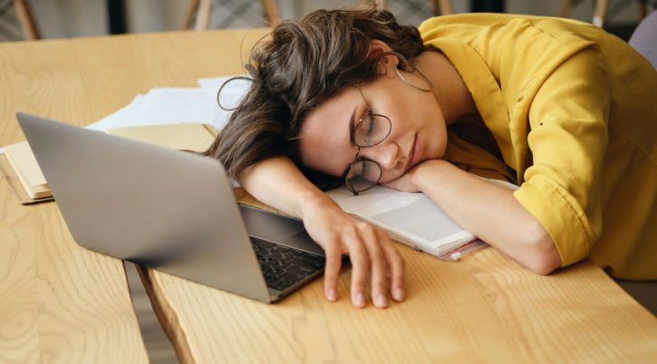 young tired woman in eyeglasses fall asleep on desk with laptop and documents under head at workplace